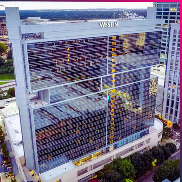 A tall modern hotel building with reflective glass windows, labeled "Westin" at the top, surrounded by trees and nearby buildings—an ideal venue reminiscent of events like Memcyco at Datos Insights FinCrime Forum 2025.