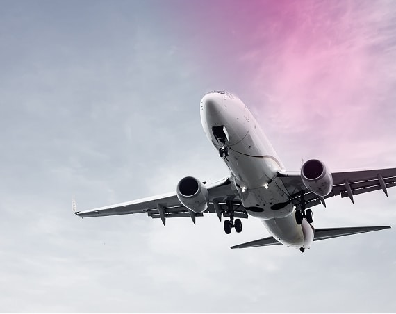A commercial airplane seen from below is banking gracefully as it flies against a cloudy sky with a pinkish hue in the upper right corner.