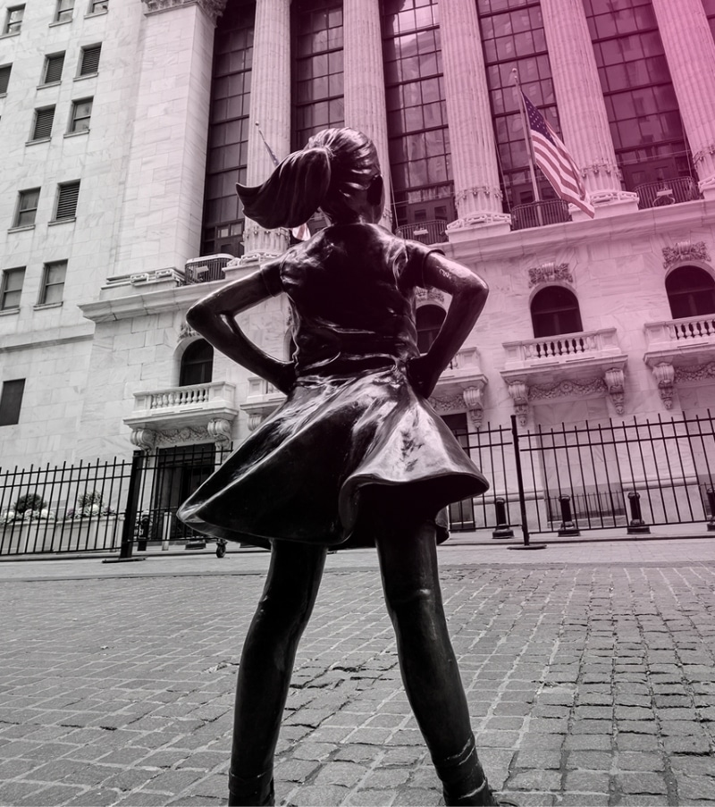 Bronze statue of a girl with hands on hips stands facing a large banking building with columns and American flags.
