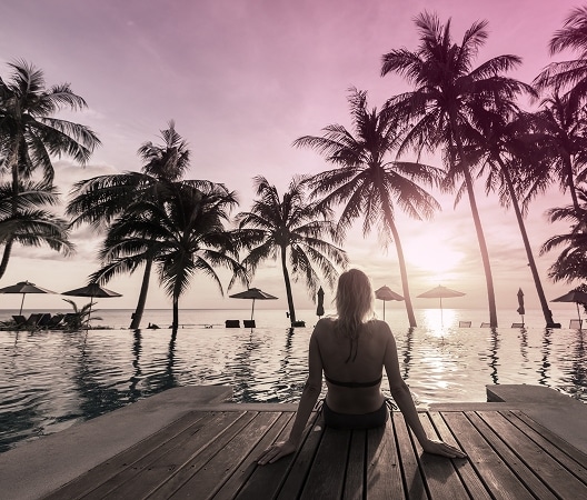 A person sits at the edge of a pool facing palm trees and a sunset sky, with lounge chairs and umbrellas in the background—an ideal setting for unwinding after a day of banking or business.