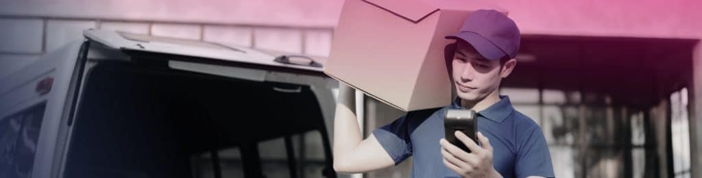 A delivery worker in a blue uniform carries a cardboard box on his shoulder and checks a handheld device near a van, likely confirming important banking documents for secure delivery.