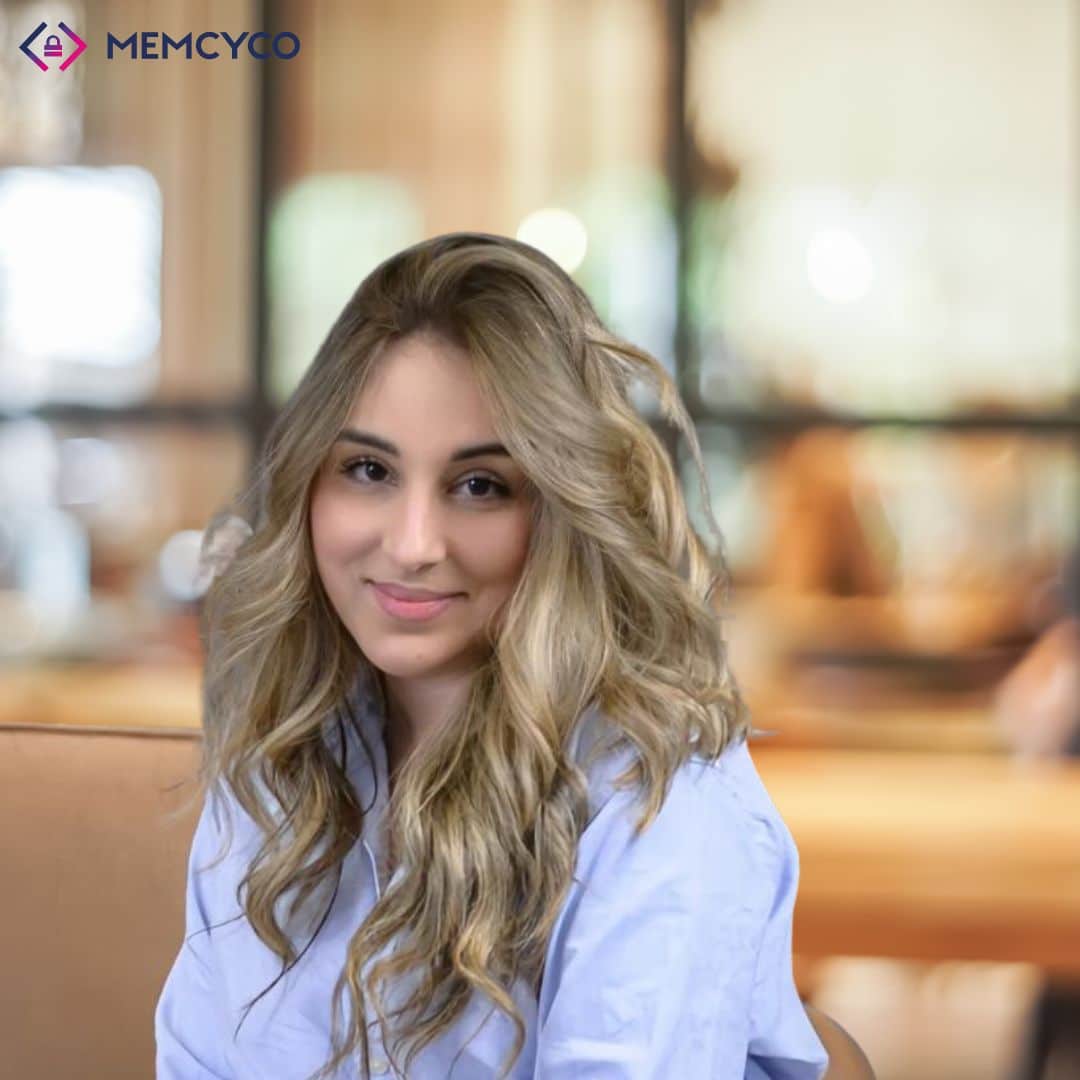 A woman with long blonde hair and a light blue shirt sits on a tan couch in a softly lit room. The logo “MEMCYCO” appears in the top left corner, highlighting Memcyco at Black Hat USA 2026.