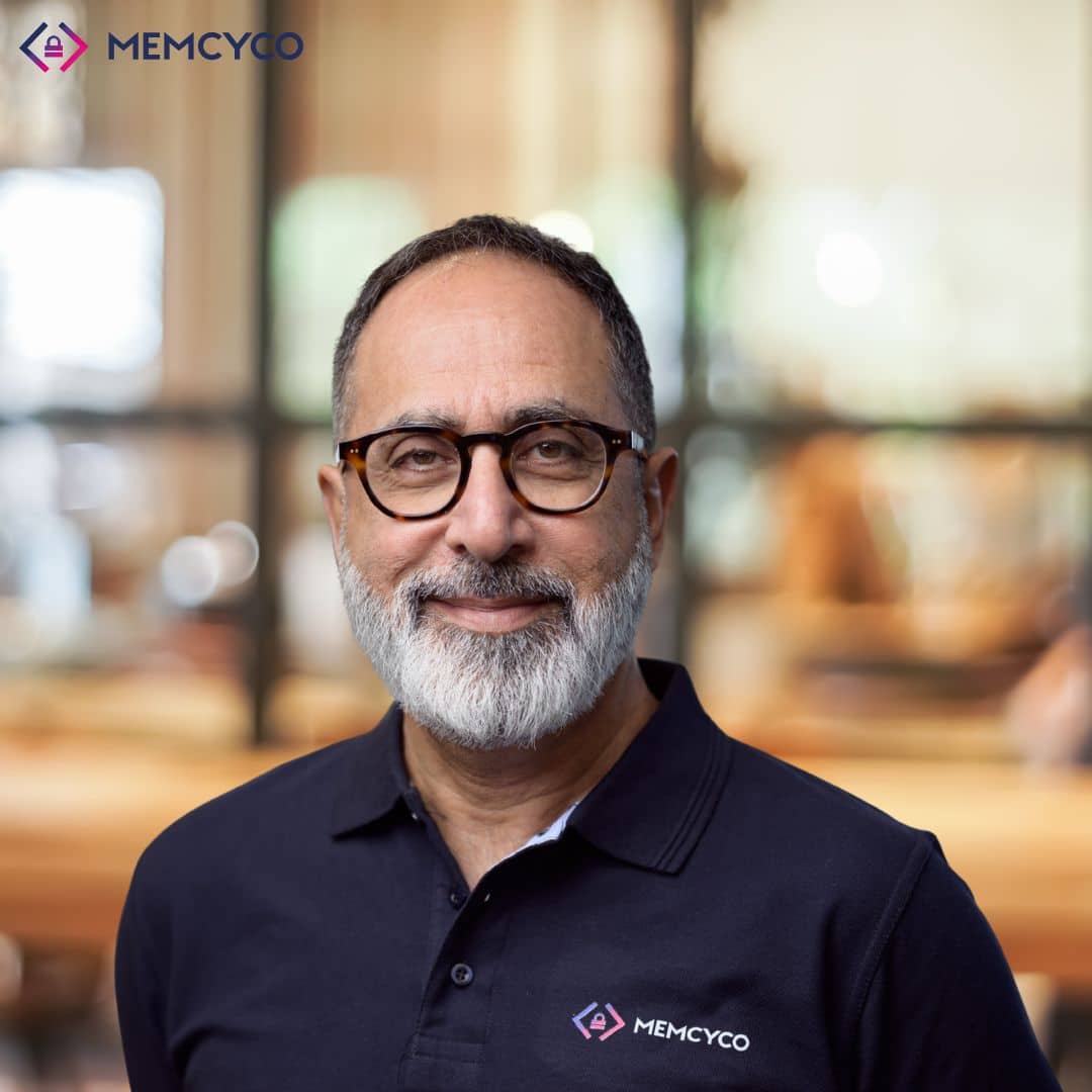 A man with glasses and a gray beard, wearing a navy MEMCYCO polo shirt, stands in a modern office with a blurred background, confidently representing Memcyco at Black Hat USA 2026.