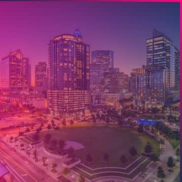 A cityscape at dusk shows tall modern buildings, a park in the foreground, and streets with some traffic under a pink and purple sky—capturing an atmosphere reminiscent of the vibrant energy that will surround Memcyco at Datos Insights FinCrime Forum 2026.
